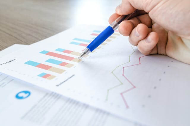 Hand with pen pointing at a bar graph and chart on paper.